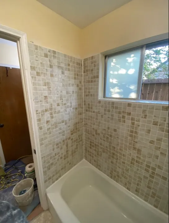 Tub-shower view with small beige mixed-tone mosaic wall tile, a white tub, a window, and an open adjoining-room doorway.