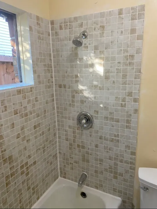 Alternate tub-shower view showing small beige mixed-tone mosaic tile, a window, exposed shower control, white tub edge, and a nearby toilet edge.