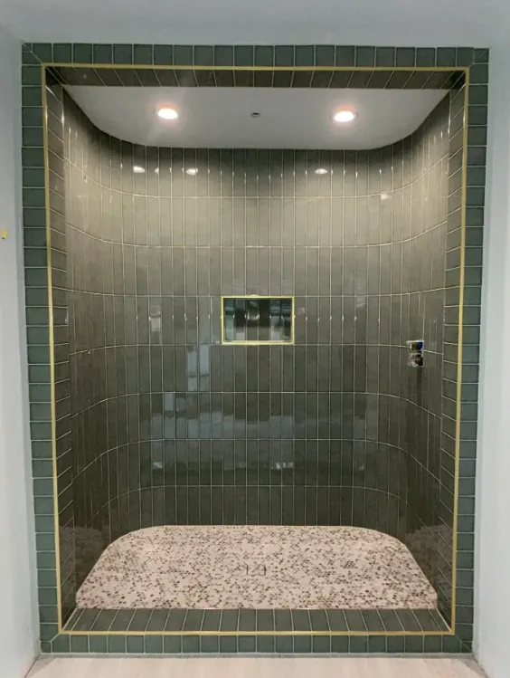 Glass-enclosed shower with narrow gray-green vertical tile, brass-toned border trim, recessed ceiling lights, and a rounded pebble-like mosaic floor.