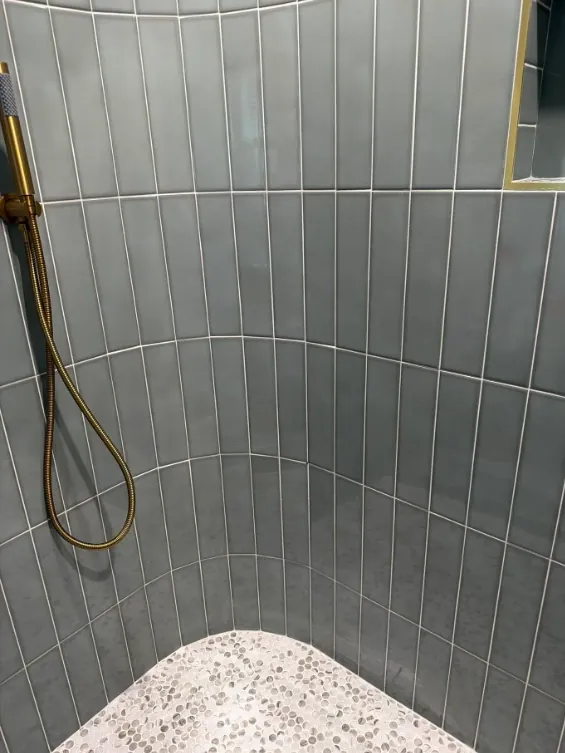 Interior view of the same shower showing curved walls of gray-green vertical tile, brass-toned handheld fixture, and rounded pebble-like floor mosaic.