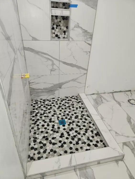 Shower-floor view with white-and-gray veined tile, a recessed niche with mixed rounded mosaic backing, and a light-and-dark rounded mosaic floor.