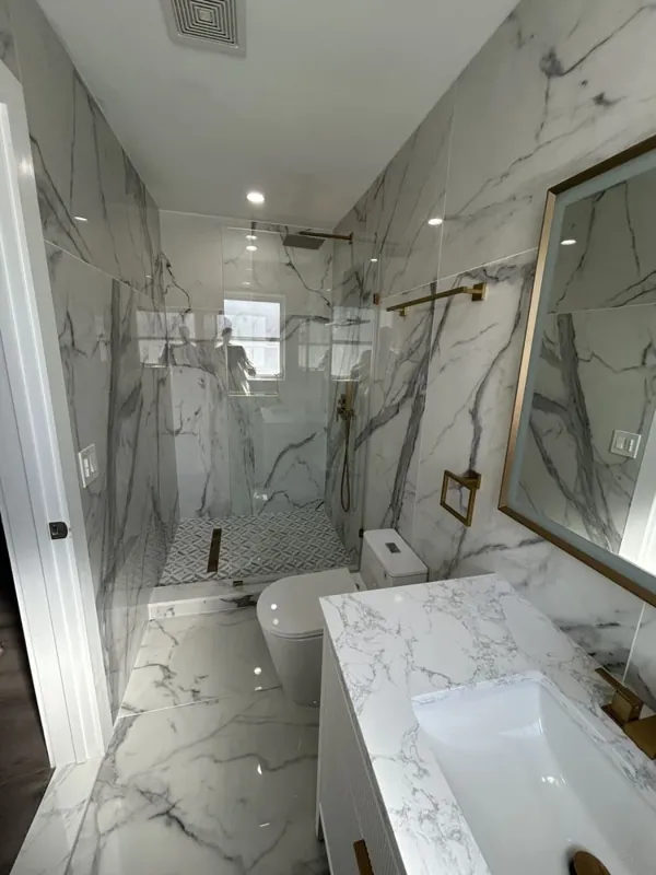 Bathroom view showing white-and-gray veined surfaces, a white vanity, clear shower glass, a toilet, warm gold-toned fixtures, and a small light shower-floor mosaic.