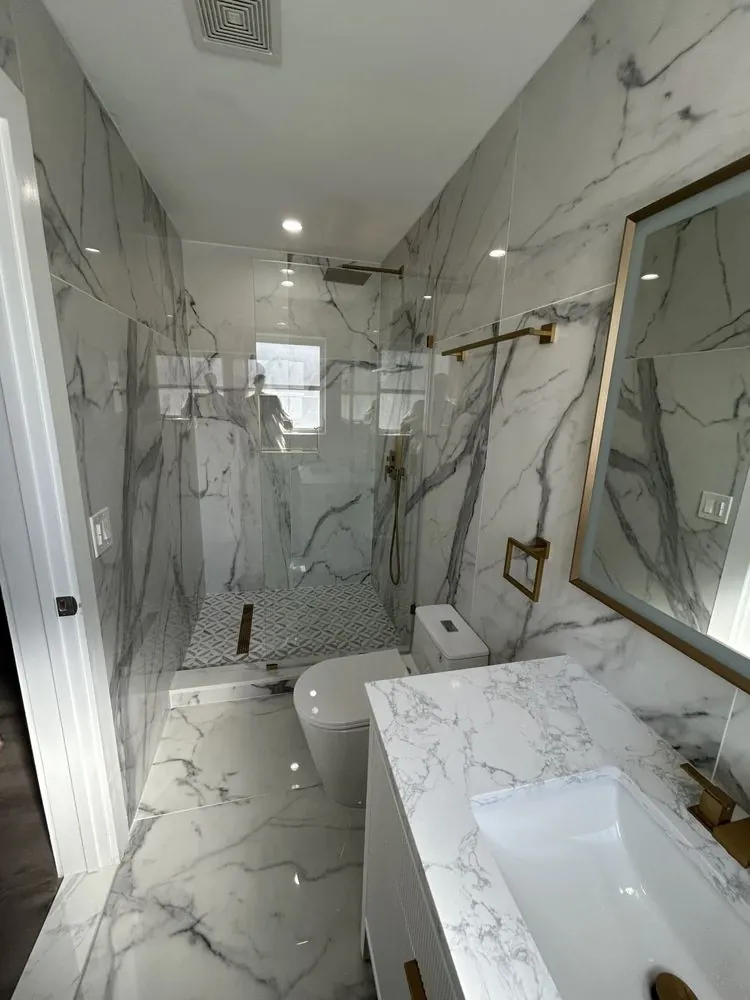 Bathroom view showing white-and-gray veined surfaces, a white vanity, clear shower glass, a toilet, warm gold-toned fixtures, and a small light shower-floor mosaic.