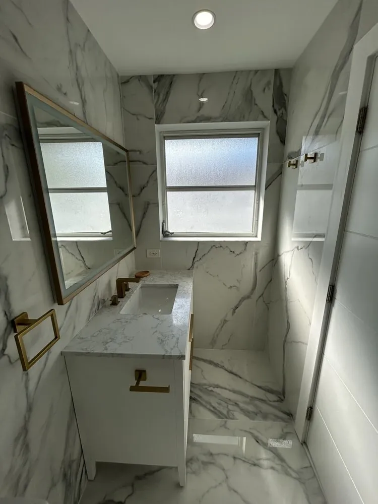 Narrow bathroom view with a white vanity, white-and-gray veined wall and floor surfaces, windows, warm gold-toned hardware, and a glass shower edge.