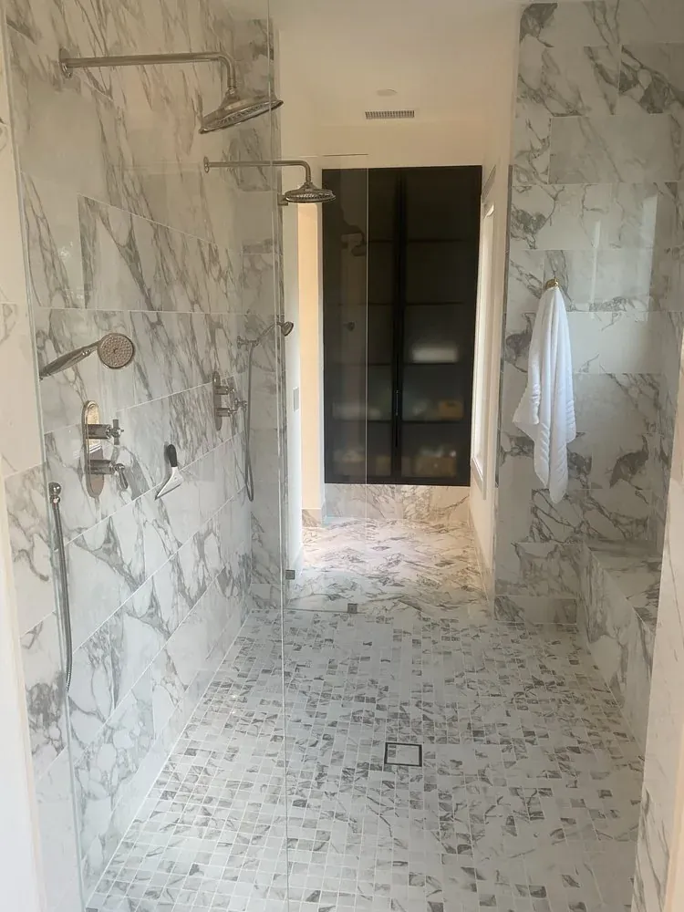 Primary-bath passage showing white-and-gray veined wall tile, a pebble-like shower floor, a glass panel, and a doorway toward a darker wet-area view.