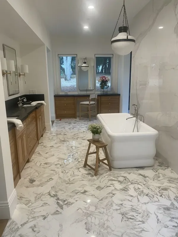 Wide primary-bath view with a freestanding white tub, wood-finish vanity, white-and-gray veined floor and wall surfaces, windows, and pendant lighting.