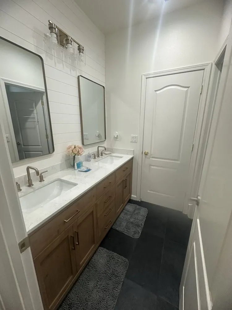 Bathroom vanity view with wood-finish double cabinetry, white counter and sinks, a large mirror, white walls, and charcoal square floor tile.