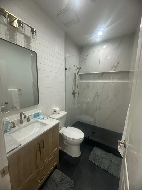 Bathroom view showing the wood-finish vanity, toilet, glass-panel shower, pale wall tile, and charcoal square floor tile.